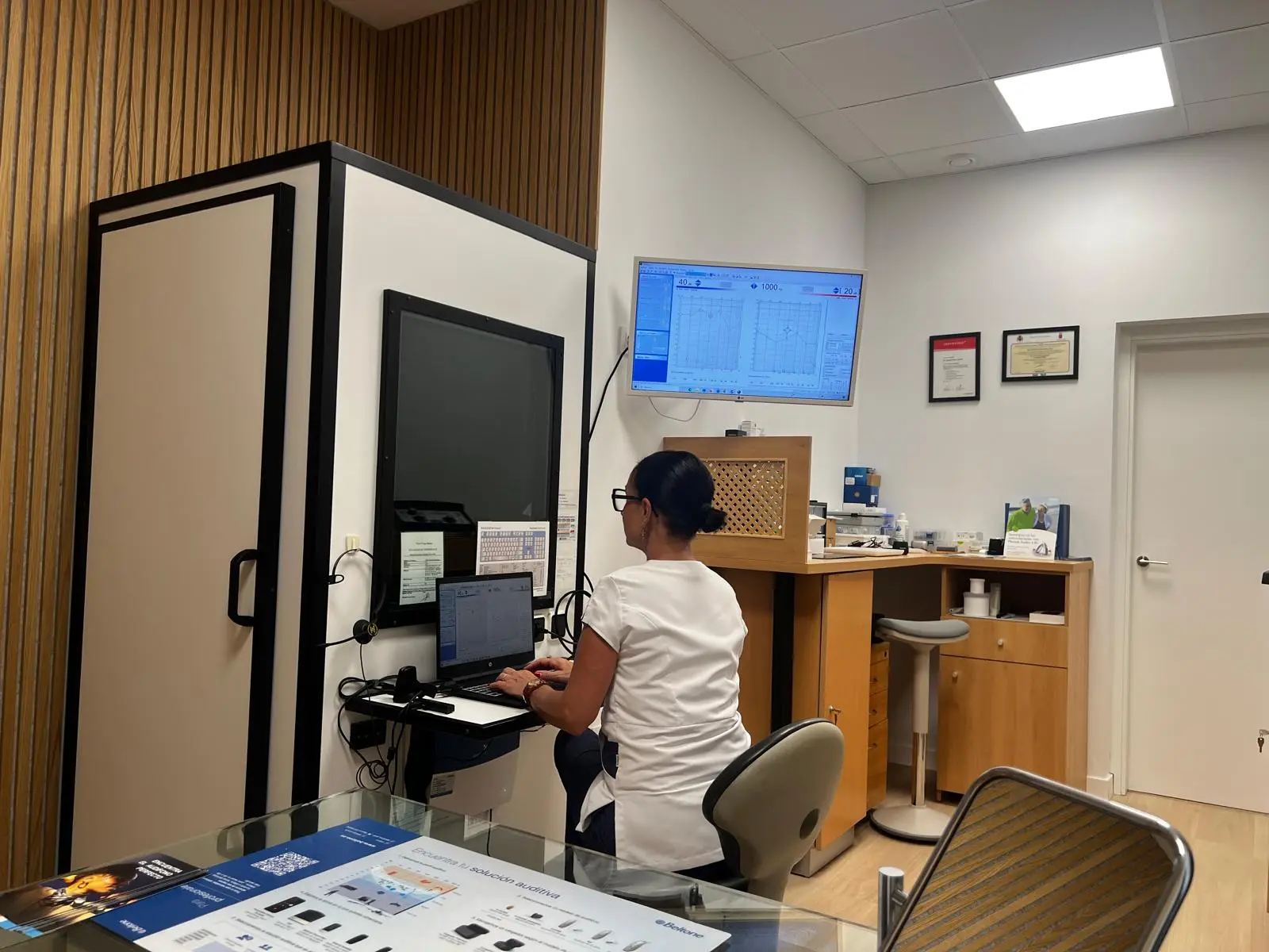Un gabinete audiológico en plena adaptación: la audioprotesista trabaja en el equipo junto a la cabina insonorizada, con un audiograma en la pantalla