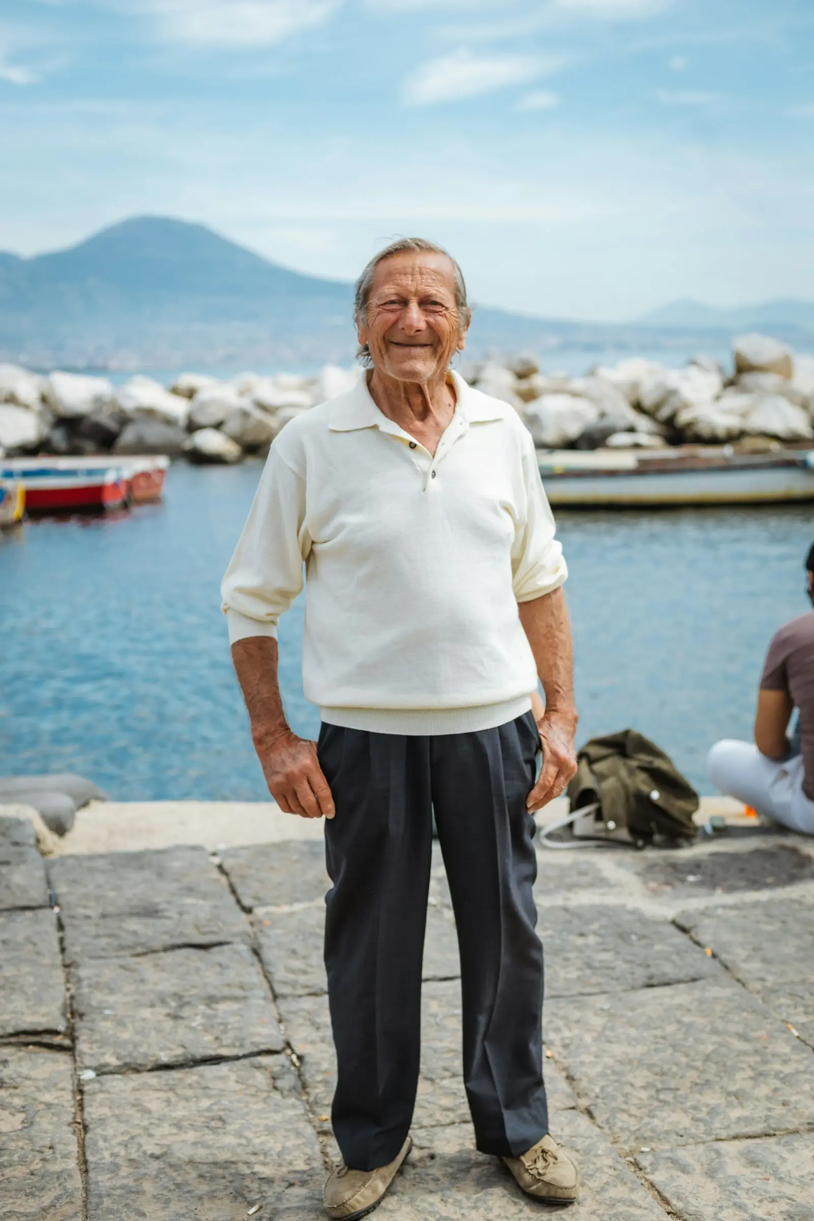 Hombre mayor de pie en el muelle de un puerto, con el mar y las barcas al fondo