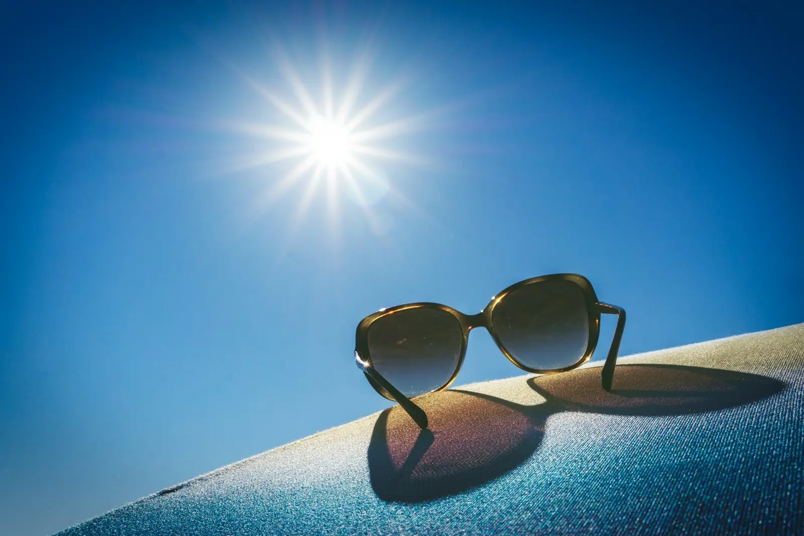 Sonnenbrille auf hellem Stoff im Gegenlicht, die Sonne steht hoch an einem wolkenlosen blauen Himmel